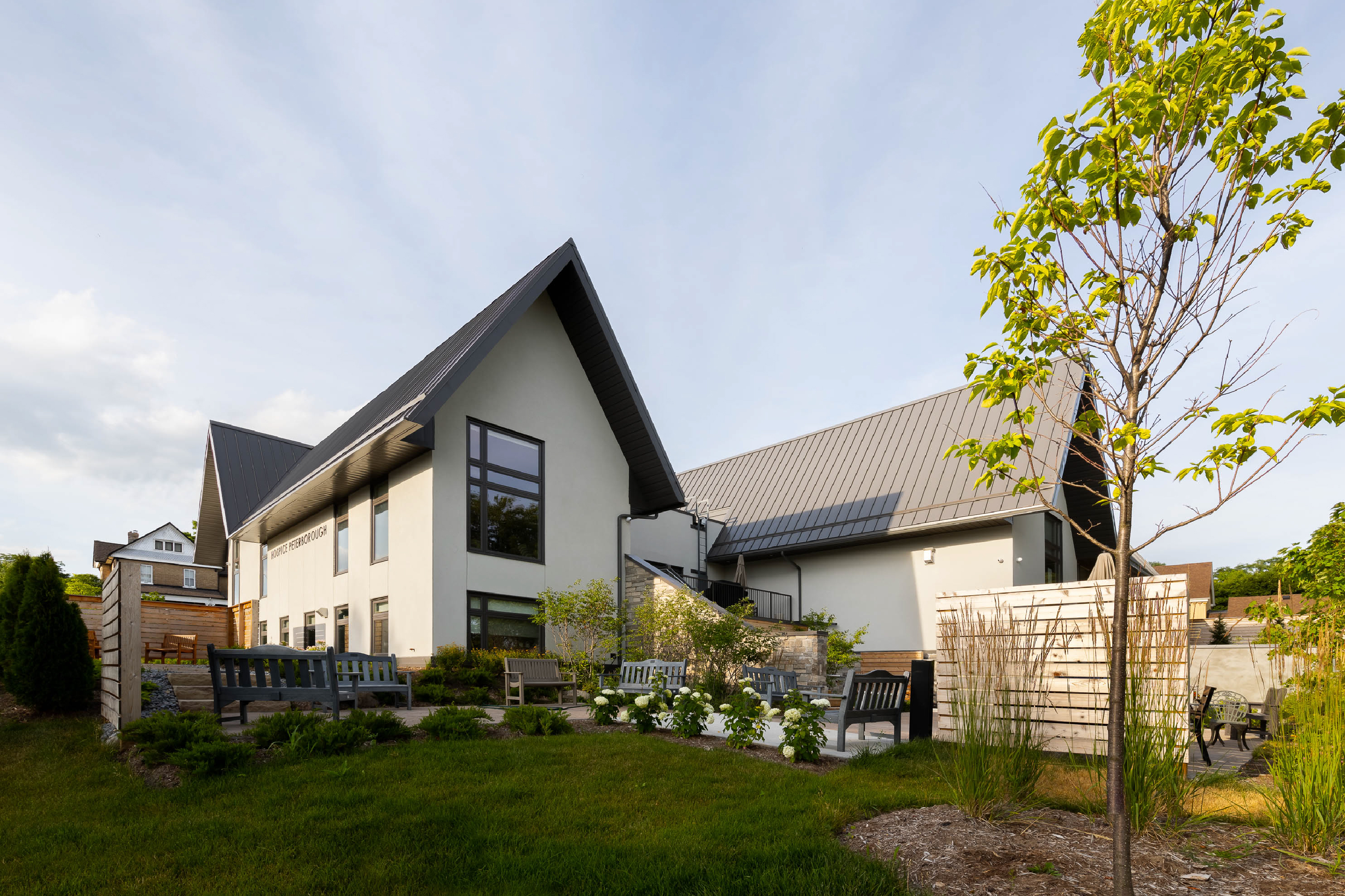 Medium-sized white stucco building with tall, steep gable roofs made of dark grey metal. In front, a garden with seating and some planting.